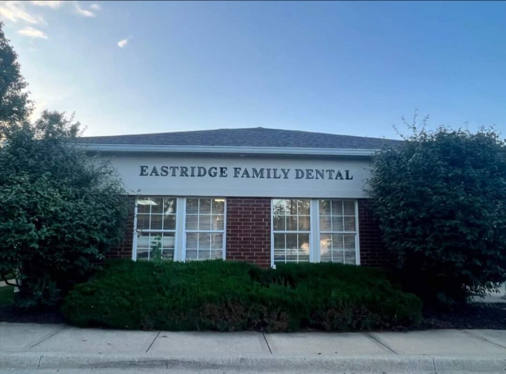 Exterior of a building with a sign that reads 'EASTRIDGE FAMILY DENTAL.' The building has a brick facade with white-framed windows, flanked by well-maintained bushes and greenery.