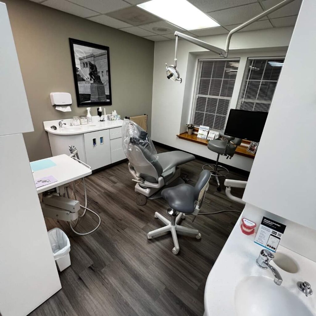 A dental examination room with a dental chair, overhead light, dental tools on a small table, a sink, a computer monitor on a desk, a stool, and cabinets. There is a window with blinds and a framed picture on the wall.